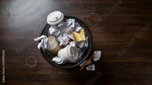 Overturned trash can with crumpled paper, paper cups, and scattered waste on wooden floor