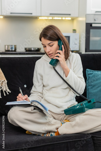 Young woman at home lifts vintage corded phone listens to voice and speaks short answer then writes notes. Caucasian girl on sofa holds handset and writes in notebook careful focus calm tone at ease