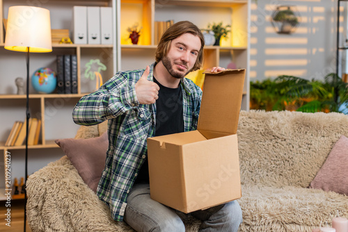 Bild auf Leinwand Young man on home sofa opens box finds and checks new purchase with keen smile