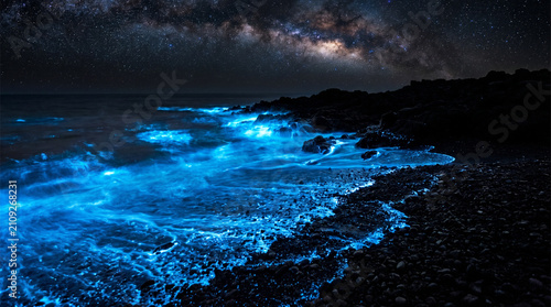 Bioluminescent glowing blue waves on rocky beach at night under Milky Way starry sky magical seascape with luminous plankton and galaxy