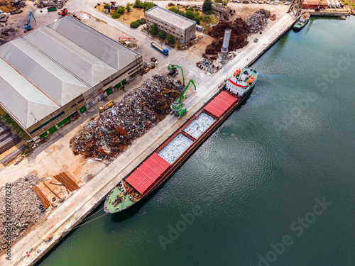 Ship docked at a warehouse with scrap metal and machinery during a sunny day in an industrial area by the water