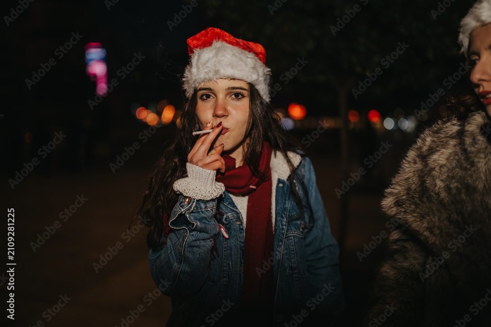 Fototapeta premium A young woman wearing a Santa hat smokes a cigarette while standing outdoors at night. She pauses during a festive evening with friends in a dimly lit street scene.