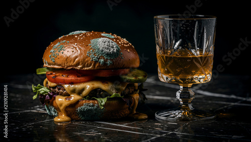 Moldy cheeseburger with green fungus sitting next to a glass of whiskey on a dark surface. Spoiled hamburger with melted cheese and sesame bun. Food safety and expiration concept for education.