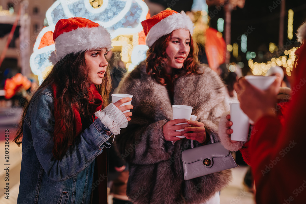 Fototapeta premium Two young women in Santa hats hold hot drinks and chat amid holiday lights. A festive night market scene with friends enjoying warm beverages and seasonal decorations.
