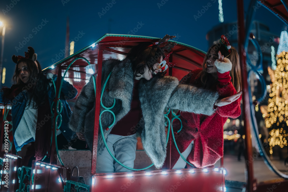 Fototapeta premium Three girls wearing festive winter clothes ride a brightly lit amusement train at night. They laugh and lean out of the carriage amid holiday lights and decorations.