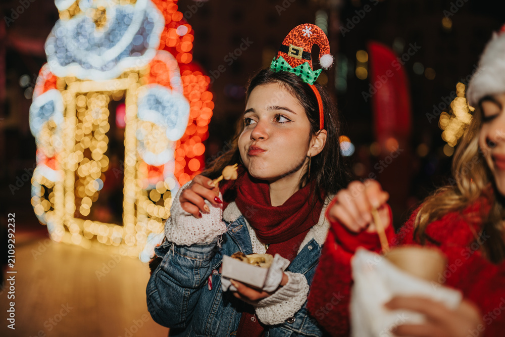 Fototapeta premium Two girls eat street food at a festive Christmas market at night. They enjoy snacks and holiday lights while standing together outdoors.