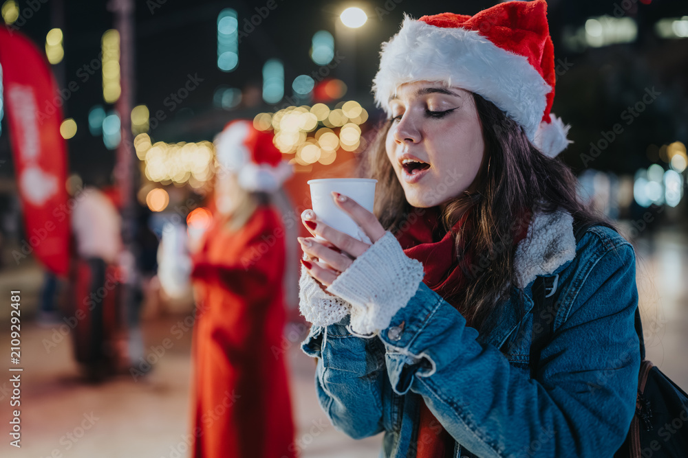 Fototapeta premium Young woman wearing a Santa hat holds a warm cup among blurred holiday lights. A girl enjoys a hot drink at a festive outdoor market at night.