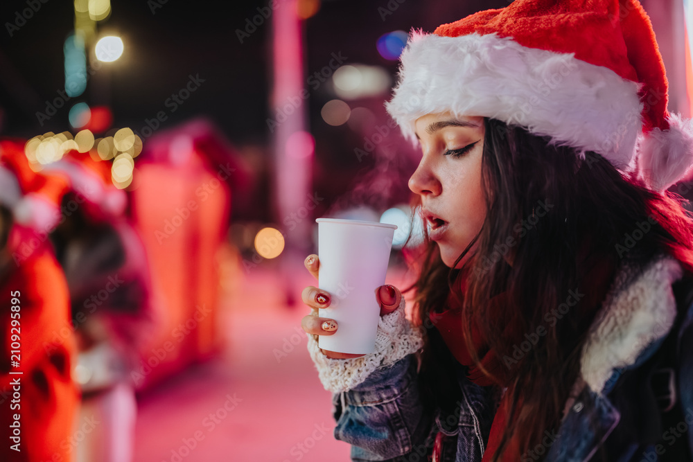 Fototapeta premium A young girl wearing a Santa hat holds a steaming paper cup while standing outdoors at night. A festive holiday market scene with Christmas lights and bokeh in the background.