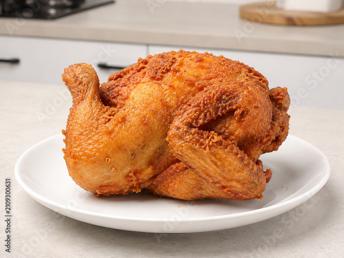 Crispy whole fried chicken with golden-brown skin served on a white plate over a modern kitchen countertop, photographed in close-up with a softly blurred kitchen background
