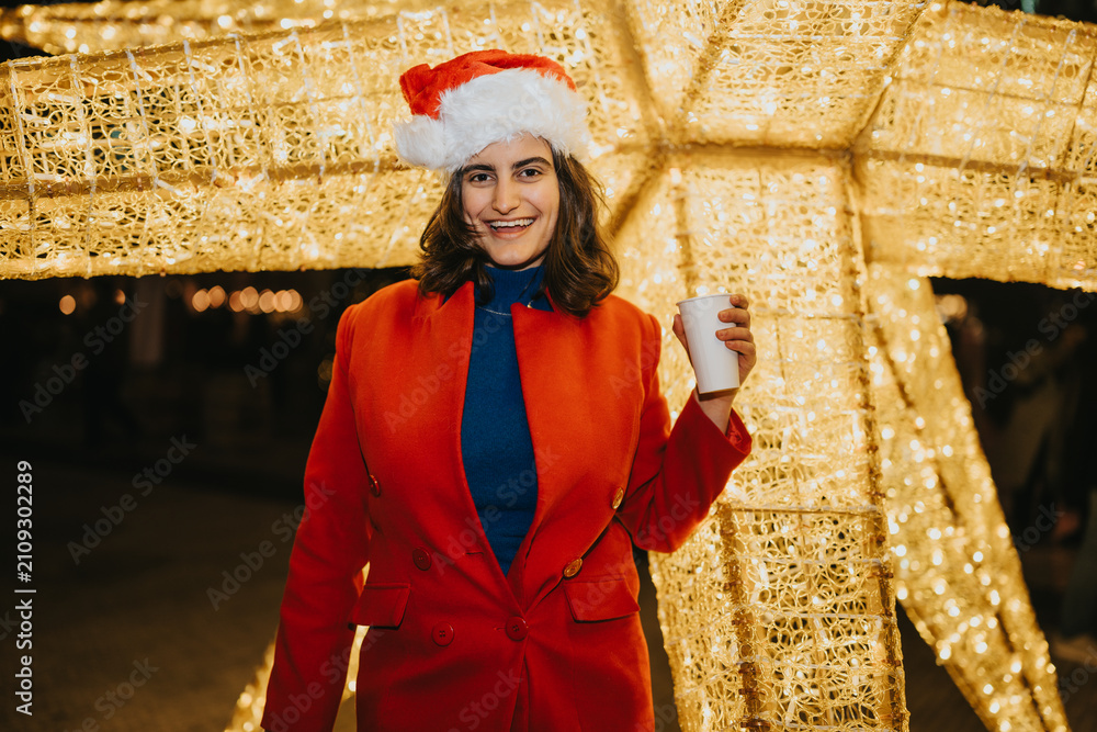 Fototapeta premium A young woman wearing a Santa hat and red coat smiles while holding a cup. She poses in front of illuminated Christmas lights at a festive outdoor market.