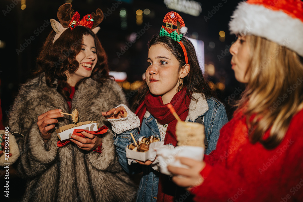 Fototapeta premium Three girls wearing Christmas hats eat street food and laugh together at night. A festive market scene showing friends sharing snacks and holiday cheer.