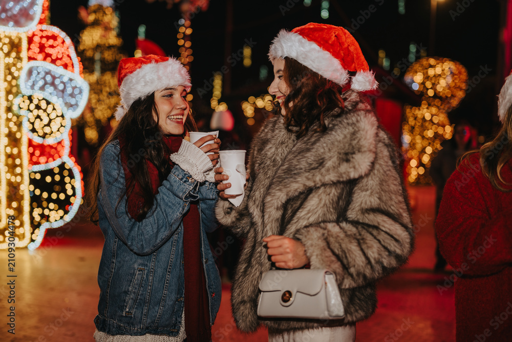 Fototapeta premium Two girls wearing Santa hats smile and hold hot drinks while chatting at a holiday market. The friends enjoy festive lights and decorations during a nighttime Christmas outing.