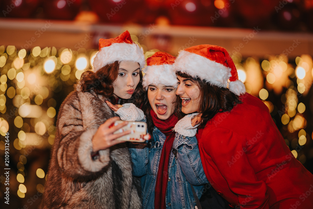 Fototapeta premium Three girls wearing Santa hats take a cheerful selfie together. The friends pose and laugh in front of warm Christmas lights at a holiday market.