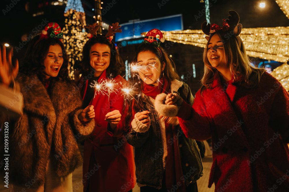 Fototapeta premium Girls hold lit sparklers and laugh while wearing festive headbands and winter coats. A joyful group of friends celebrates outdoors at night surrounded by holiday lights.
