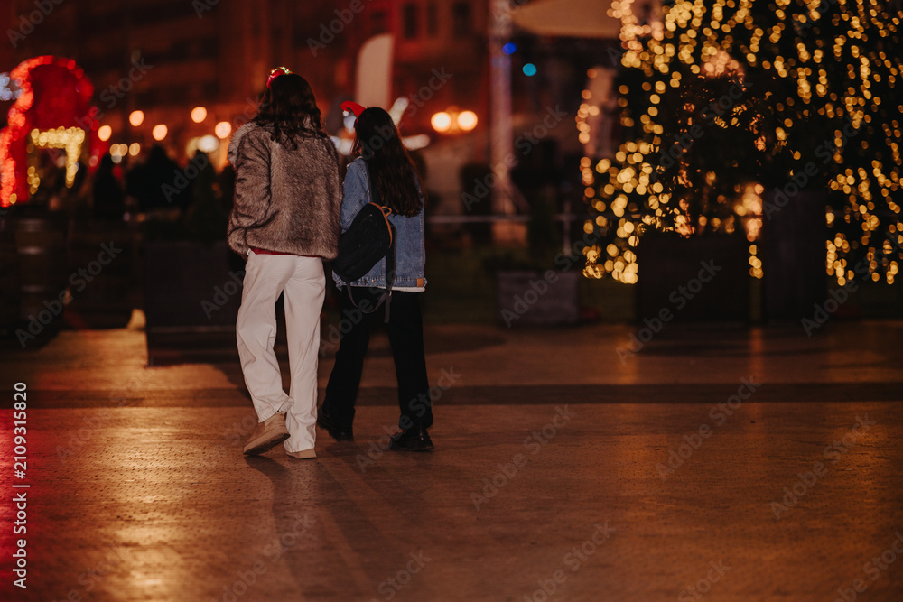 Fototapeta premium Two girls walk together at night next to illuminated holiday decorations. They stroll through a city square with warm festive lights in the background.