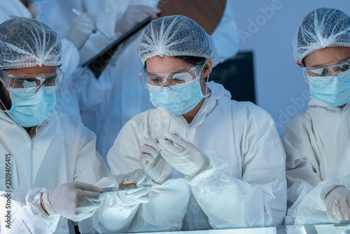 Three specialists in white protective suits and masks studying small samples on an illuminated table, semiconductor inspection in cleanroom facility.