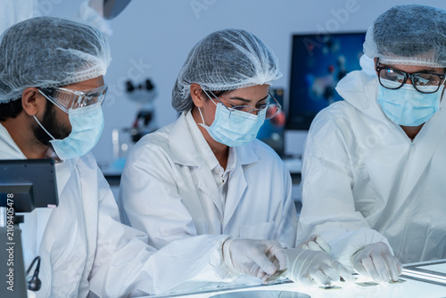 Three specialists in protective suits and masks examining material specimens on an illuminated table, semiconductor production in blue-lit cleanroom.