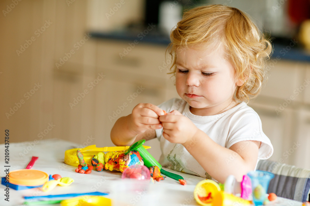 Obraz premium Adorable cute little toddler girl with colorful clay at home. Healthy baby playing and creating toys from play dough at nursery or playschool. Small kid molding modeling clay, creative activity