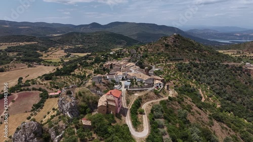Flying across a scenic Spanish village in the Pyrenees, located on the top of a rocky mountain. Wide shot including surrounding landscape.