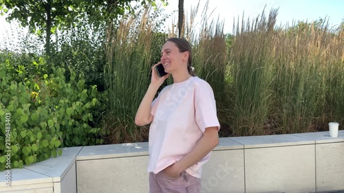 Young woman in pink t-shirt talking on smartphone and smiling outdoors near concrete bench with grasses background. communication, lifestyle, technology, outdoors, happy