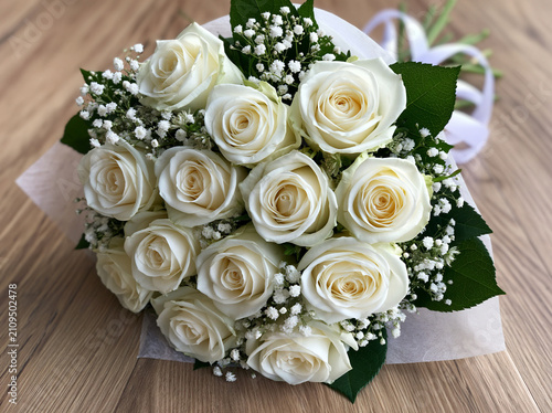 A beautiful bouquet of fresh white roses with delicate babys breath flowers and green leaves, wrapped in elegant paper, resting on a rustic wooden surface