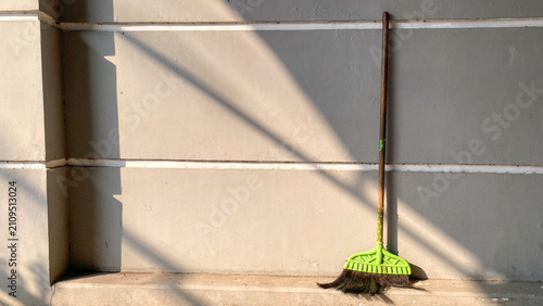 Traditional broom leaning against a gray concrete wall illuminated by warm natural sunlight. Minimal outdoor cleaning concept featuring shadows, textured wall, and copy space.