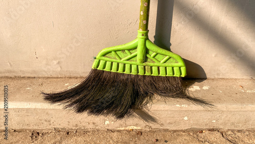 Close-up of a traditional broom resting against a gray concrete wall with warm sunlight. Detailed view of the broom bristles and textured outdoor surface, representing household cleaning and maintenan