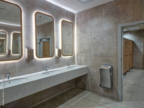 Modern public restroom interior with illuminated mirrors, white counter sinks, and grey tile walls.