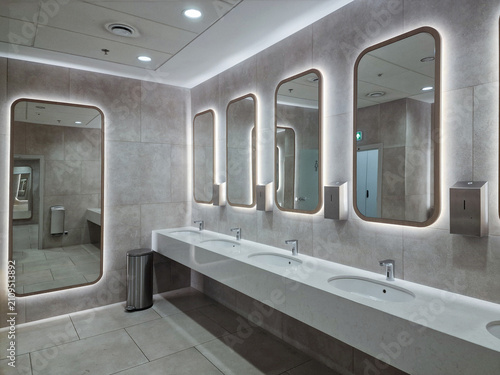 Modern public restroom interior with illuminated mirrors, white counter sinks, and grey tile walls.