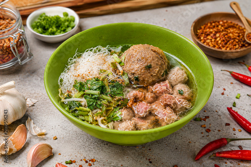 Traditional Indonesian bakso soup served in a green bowl with a large tendon meatball, small beef meatballs, diced beef, beef trimmings, rice vermicelli, fresh mustard greens, and bean sprouts in a sa