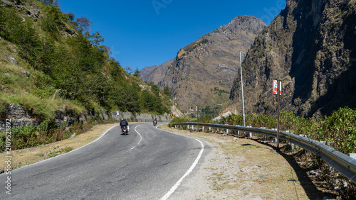 Two motorcyclists riding on a winding road through a mountainous landscape with lush greenery and clear blue skies in Joshimath near Badrinath