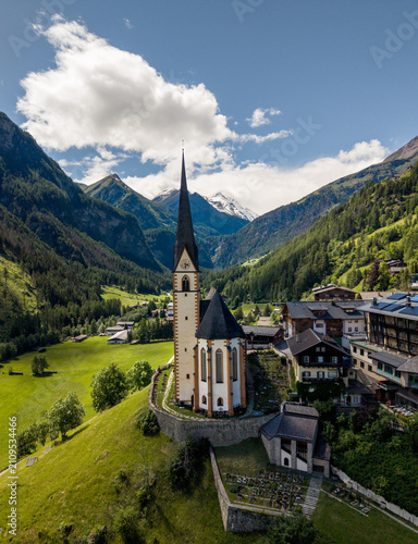 Hof, Austria - June 11 2026: Aerial drone footage of Pfarrkirche Heiligenblut, church in Hof, Austria. Beautiful weather with snowy mountains in the background. Heaven in the Austrian Alps. Grossglock