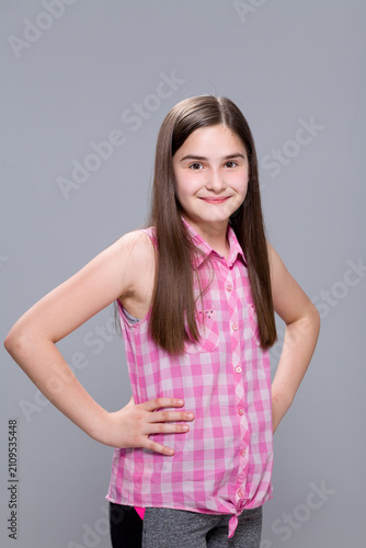 Studio portrait of a confident smiling young girl in a pink plaid sleeveless shirt with hands on hips against a grey background.