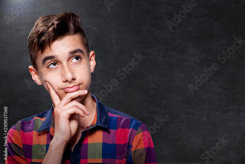 Studio portrait of a thoughtful teenage boy in a colorful plaid shirt looking up with hand on chin against a dark textured background with copy space.