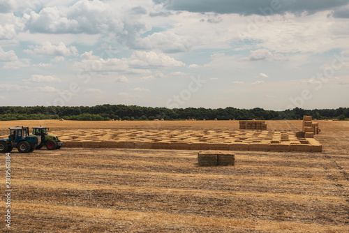 Tractors on vast agricultural field of golden wheat hay bales with cloudy sky