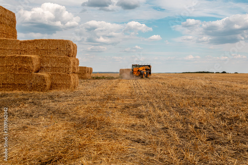Tractor moves hay bales across golden field under  cloudy sky. Rural agriculture scenery of seasonal harvest works