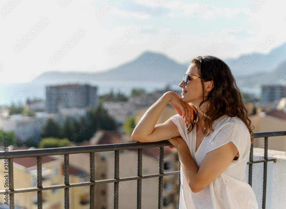 Obraz premium Beautiful young woman standing on apartment balcony and enjoying sunny summer day. City view background.