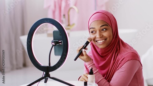 Smiling Young Muslim Woman With Light Skin Applying Makeup With Brush In Front Of Ring Light At Home. She Is Enjoying Her Beauty Routine While Looking Confident And Happy.