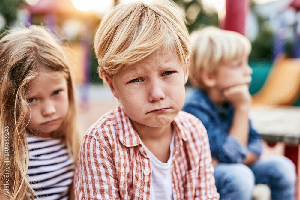 Fototapeta premium Three young children with sad expressions sitting outdoors, one boy looking directly at the camera with disappointment.