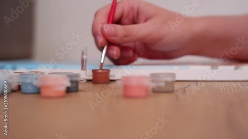 Artist's hand dipping a paintbrush in brown acrylic paint and then drawing on a numbered canvas