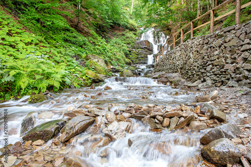 Water flows down rocks in a stream with a waterfall in a forested area on a sunny day
