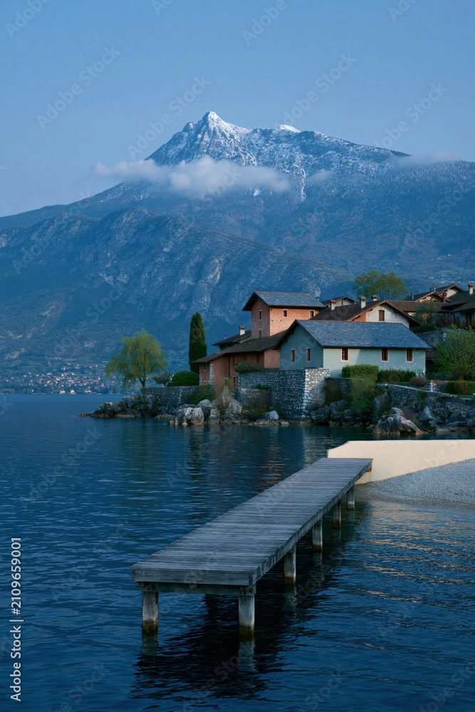 Fototapeta premium Tranquil lakeside village with wooden dock and snowcapped mountain backdrop for serene wallpaper and banner design