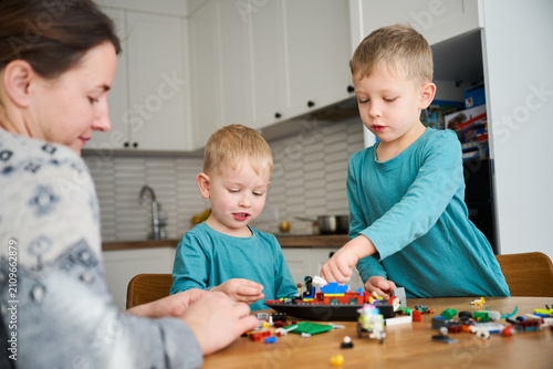 Mother and two young boys building toy model from colorful construction blocks at table. Focused family activity, creativity and learning together at home