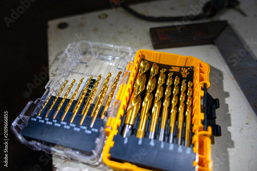 Golden drill bit set in plastic case on workbench with warm light and shallow depth of field showing organized metal tools and workshop atmosphere