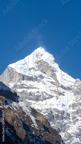 A majestic snow-capped mountain peak under a clear blue sky with rocky terrain and patches of snow in the foreground, framed by rugged cliffs and distant peaks, bathed in sunlight