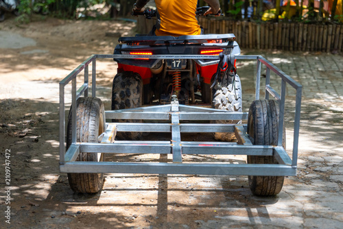 Utility trailer frame hitched to off road ATV with rider in yellow shirt, dusty pathway and rural outdoor setting showing metal construction and spare tire emotion of readiness