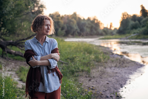 Schilderij op canvas miling woman wrapped in a shawl standing by a peaceful river at sunset, enjoying a quiet moment in nature