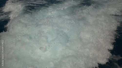 Turbulent white foam water trail behind a large boat in a calm blue sea on a sunny day, slow motion