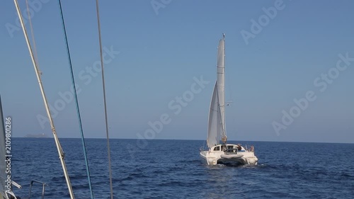 Point of view shot from a sailboat looking at a modern luxury catamaran cruising under full sail on the deep blue calm waters of the Mediterranean Sea. The scene captures the beauty of nautical travel
