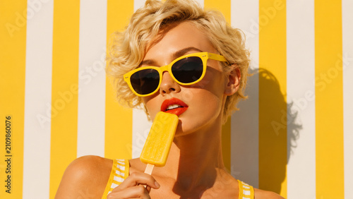 A stylish blonde woman in yellow sunglasses enjoys a popsicle against a striped background.